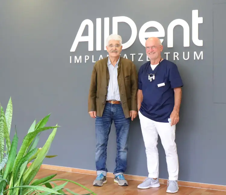 Zwei M&auml;nner stehen l&auml;chelnd vor einer Wand mit der Aufschrift AllDent Implantatzentrum.