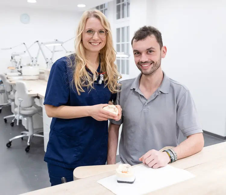 Eine Frau in blauer Arbeitskleidung steht neben einem Mann in grauem Poloshirt an einem Tisch. Beide l&auml;cheln in die Kamera, die Frau h&auml;lt ein Zahnmodell in den H&auml;nden, auf dem Tisch liegt ein weiteres Modell.
