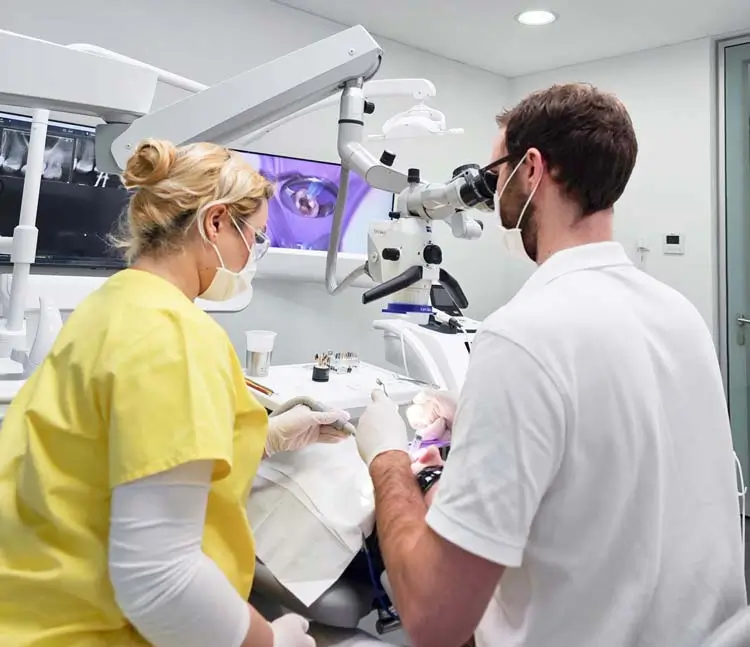 Zahnarzt mit Mundschutz behandelt einen Patienten. Einer arbeitet mit einem Mikroskop, w&auml;hrend die andere Person assistiert. Im Hintergrund sind Monitore mit Zahnaufnahmen zu sehen