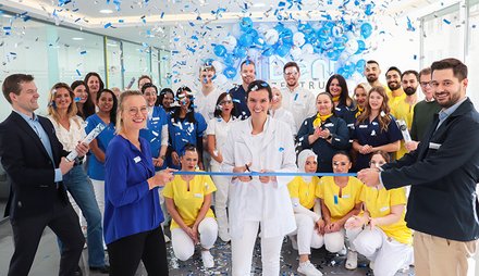 Menschen in bunter Arbeitskleidung schneiden ein blaues Band durch, während blaue Luftballons und Konfetti fallen.