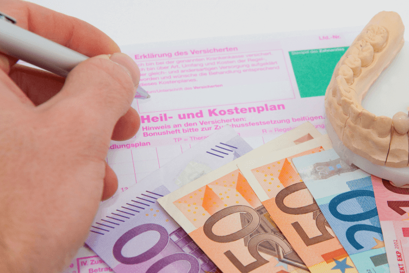 Hand schreibt auf Heil- und Kostenplan, darunter liegen verschiedene Geldscheine. Seitlich liegt ein Modellgebiss